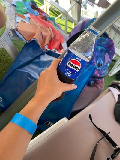 A person's left arm, wearing a bright blue wristband, is in the foreground, holding a plastic bottle of Pepsi. The bottle is mostly full of dark liquid, and the Pepsi logo is clearly visible. Behind the arm, there is a large blue reusable shopping bag with "Scandinav..." printed on it. To the left of the bag is a colorful beach towel with cartoon characters. In the background, there is a metal railing, a purple backpack with a Stitch character on it, and lounge chairs. The environment appears to be outdoors, possibly at a resort or beach, given the lounge chairs and beach towel. The ground appears to be covered in artificial turf. There is no discernible connection to the city of Zazymia, Ukraine, in the provided image.
