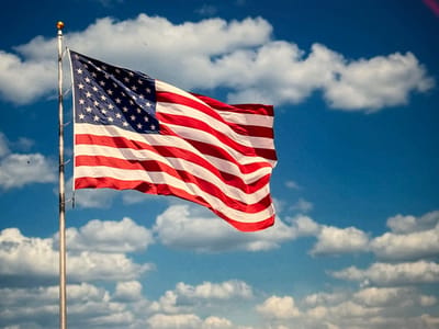 A vibrant American flag, prominently displayed on a metal pole, waves in the wind against a bright blue sky. Puffy white clouds scatter across the background, enhancing the image's patriotic feel. The flag's red and white stripes and star-studded blue field are clearly visible, adding to its symbolic significance.