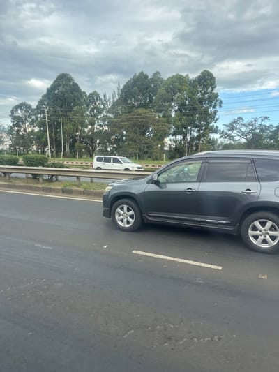A gray SUV is parked on an asphalt road. A white van is visible behind the SUV, and both are adjacent to a concrete barrier. Beyond the barrier, lush green trees stretch towards a cloudy sky with visible power lines. A white lane marking is painted on the road's surface.