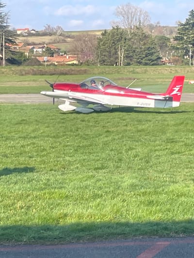 At Aérodrome de Vienne Reventin (LFHR) in Chonas-l'Amballan, France, a red and white single-engine aircraft, tail registration F-JVOV, is situated on a patch of green grass adjacent to an asphalt surface. The aircraft features a visible propeller, a transparent bubble canopy, and white wheel fairings. A white "Z" emblem marks the red vertical stabilizer. Two occupants, wearing headsets, are seated within the cockpit: one in the left pilot's position and one in the right. The immediate environment consists of sunlit green grass with an asphalt edge. In the background, a landscape of rolling hills contains scattered residential structures with red-tiled roofs and diverse tree cover. The sky is light blue with sparse cloud cover.