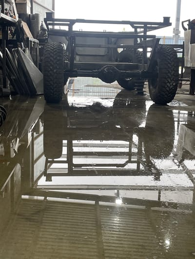 This image shows the underside of a vehicle frame, possibly a truck or tractor, with large tires visible. The vehicle appears to be in some sort of industrial or workshop setting, with metal structures and other components in the background. The ground is wet and reflective, suggesting it might be raining or the area has recently been cleaned. The lighting suggests it is daytime. There is no visible text in the image. The scene is primarily focused on the mechanical parts of the vehicle.