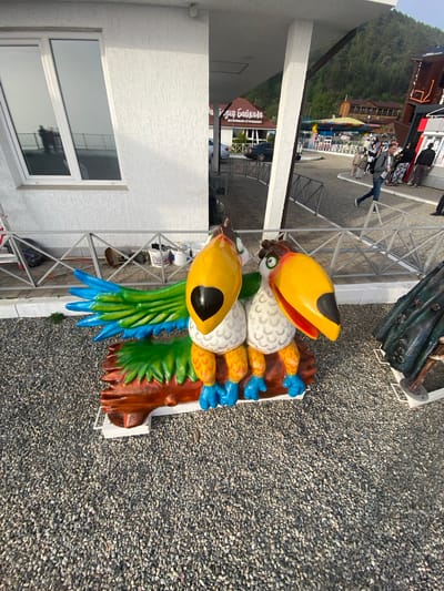 Two brightly colored toucan statues are the primary subjects in the foreground of this image, positioned on a gravel surface. The statues are detailed with yellow beaks, white bodies, and colorful wings and feet, with one appearing slightly larger than the other. To the right of the toucans, a dark, textured object, possibly another statue or decoration, is partially visible. In the background, a white building with a reflective window is present, showing a reflection of people and a railing. Beyond the building, a street scene unfolds with several people walking, cars, and amusement park attractions, including a carousel-like structure. A sign with Russian text, "для Байкала," which translates to "for Baikal," indicates a location related to Lake Baikal. The environment appears to be an outdoor tourist area, likely in the town of Listvyanka, Russia, a popular destination on the shores of Lake Baikal. The lighting suggests it is daytime.