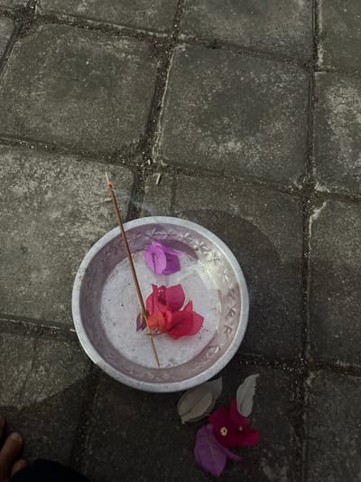 An overhead shot shows a small, round, metallic dish containing two bright pink bougainvillea petals and one purple bougainvillea petal. A single incense stick, lit and emitting a thin wisp of smoke, is placed in the dish. The dish is positioned on a stone patio with rectangular pavers. Several more bougainvillea petals, some pink and some light gray, are scattered on the patio stones to the right of the dish. A person's bare foot is visible in the lower left corner of the frame. The scene is likely in the city of Mendoyo, Indonesia, based on the presence of bougainvillea and the common practice of offering incense and flowers.