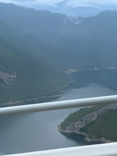The image depicts a scenic view of a reservoir and surrounding mountains, likely from a vantage point within or overlooking the town of Liaskovo, Bulgaria. In the foreground, a white railing runs horizontally across the lower portion of the frame, suggesting the viewer is on a bridge, balcony, or overlook.

The middle ground is dominated by a body of dark, calm water, a reservoir, which curves into the distance. The water's surface reflects the muted tones of the sky and the surrounding landscape. Along the edge of the reservoir, the terrain slopes steeply upwards, covered in dense, dark green coniferous forest. Rocky outcrops and sheer cliffs are visible on some of these slopes, particularly on a prominent point of land jutting into the water in the lower right quadrant.

The background consists of layers of mountains, their peaks shrouded in a hazy, bluish-grey mist, indicative of a cool or overcast day, possibly early morning or late afternoon. The distant mountains are softened by the atmospheric conditions, creating a sense of depth and vastness. A small, isolated building with a light-colored roof can be spotted nestled on a hillside overlooking the reservoir in the middle distance. The overall impression is one of natural tranquility and rugged mountainous beauty.