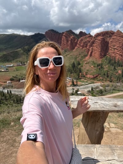 An outdoor selfie shows a woman posing in the scenic mountainous region of Jeti-Ögüz, Kyrgyzstan. The image is taken from a slightly elevated perspective, looking down into a valley. The woman, with long blonde hair, wears large white square-framed sunglasses and a light pink t-shirt. On her left sleeve, a small black tag features a white, cartoon-like panda or bear face. The text "PROD" is partially visible on the front of her shirt. She also wears a gold hoop earring on her left ear. A grey crocheted or woven bag rests on a rustic wooden railing beside her.
In the background, the dramatic landscape features towering, textured red rock formations, characteristic of the Jeti-Ögüz area, partially covered with green vegetation. Below the red rocks, a valley extends, dotted with trees, green fields, and several small buildings, suggesting a village or scattered settlements. A winding road or path is also visible through the valley.
The sky is partly cloudy with patches of blue. The bright lighting suggests it is daytime, likely late morning or early afternoon, with clear shadows cast by the wooden railing. The woman maintains a neutral expression as she captures the moment against the striking natural backdrop.
