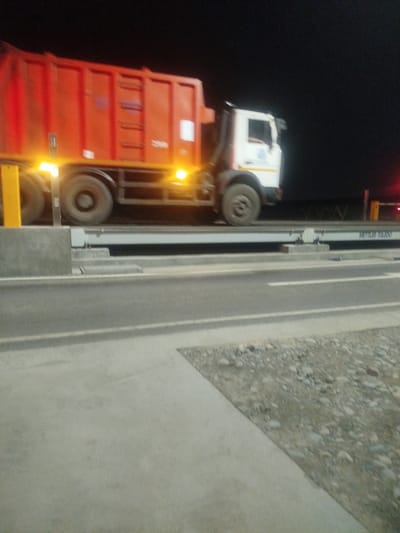 The image shows an orange garbage truck with a white cab stopped on a weighbridge at night. The truck is positioned on a raised platform with yellow markings. The background is dark, suggesting it is nighttime. The weighbridge has a metallic surface and appears to be part of a road infrastructure, possibly a toll station or a weight check point. The location is identified as Түндүк айланып өтүүчү жол 84 га, Kyrgyzstan. The truck's tires and undercarriage are visible. There are lights on the truck and the weighbridge infrastructure. The overall scene is industrial and functional, related to transportation and logistics.