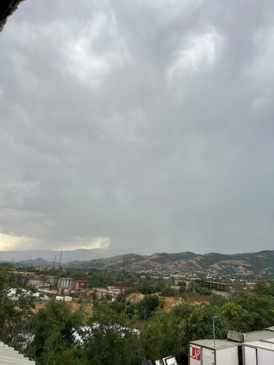 The image depicts a landscape under a cloudy sky, likely taken from an elevated vantage point. In the foreground, there is a white structure that appears to be the roof of a building, with some greenery visible below it. Partially visible in the lower right corner are the tops of white containers, one of which has a red "JP" logo.
The midground features a collection of large, cylindrical industrial tanks, some of which appear weathered and rusted. Beyond the tanks, there is a landscape with dry, rolling hills dotted with trees and some buildings, suggesting a semi-rural or industrial area. Several tall, thin structures resembling towers or antennas are scattered across the scene. The background is dominated by a range of hills and mountains, also covered in vegetation, and shrouded in the overcast conditions. The overall atmosphere is somber due to the heavy cloud cover.
While the image shows an industrial area with tanks and the landscape of hills, there is no specific visual information within the image that definitively places it in the city of Struma, Bulgaria. The description is solely based on the visual elements present in the photograph.
