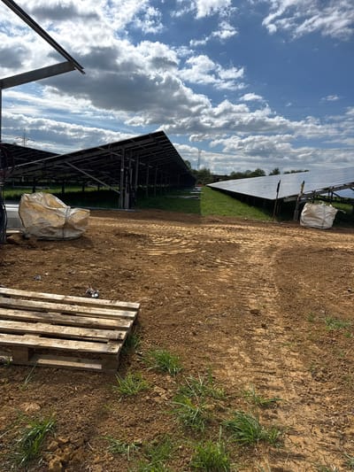 The image depicts a construction site in Beanacre, United Kingdom, likely for a solar panel installation. Rows of solar panels, some fully installed and others under construction, stretch across the landscape under a partly cloudy sky. In the foreground, there is a dirt and muddy ground, with a wooden pallet and a large white bag lying on the surface. Scattered patches of green grass are visible. Metal frames and electrical wiring are also apparent in the view, adding to the construction site context.