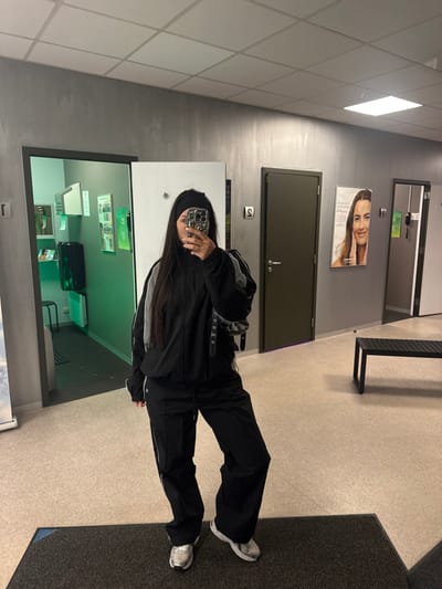 The image displays a full-length mirror selfie of a female individual inside an indoor facility in Temse, Belgium, identified as likely an aesthetic clinic or beauty center. The subject, an adult female with long dark hair and a black headband, is dressed in a black tracksuit featuring grey/white stripes on the sleeves, paired with silver athletic shoes. She holds a smartphone with a patterned case with both hands, obscuring her face above the nose, while holding a small, black, studded shoulder bag on her left side.
The room features grey, textured walls and a light-colored floor, with a dark grey mat positioned in the immediate foreground. The ceiling is a white grid with recessed square lighting panels. Three doorways are visible:
1. On the far left, a doorway leads into a room illuminated with a bright green/cyan light, containing a black waste bin, a white stool, and several white panels mounted on the wall. A "T" symbol is affixed to the wall next to this doorway.
2. In the center-right, a solid dark brown door is present, featuring a silver handle. A vertical "S" symbol is attached to the wall beside this door.
3. On the far right, a third doorway is partially visible.
A black slatted bench is positioned against the right wall. Between the two rightmost doors, a vertical poster advertises aesthetic services, displaying a woman's face and text mentioning "natuurlijk mooi" (naturally beautiful), "huidverjonging" (skin rejuvenation), "fillers," and "botox."