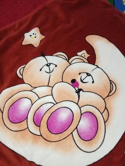 A dark red, textured fabric, consistent with a blanket, is depicted in Ciputri, Indonesia. The fabric features an illustration of two light brown cartoon teddy bears embracing. The bear positioned on the left has a smiling expression with closed eyes and eyelashes. The bear on the right has closed eyes and a pink object in its mouth. Both bears exhibit pink inner ear details and pink paw pads. A white crescent moon shape is partially visible behind the bears to the right. Two small, light brown cartoon stars with smiling faces are present in the background, one above the left bear and a smaller one near the right bear's head.