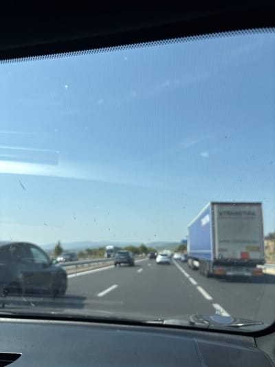 The image shows a view from the windshield of a car driving on a highway. The sky is blue with some wispy clouds. There are several cars and a large blue and white truck on the road. The highway appears to be in a rural or semi-rural area, with hills visible in the distance. The car is in motion, as indicated by the blurred foreground and the perspective of the road. There are some smudges and dust on the windshield. There is no indication of the location being specifically within the city of Bages, France.