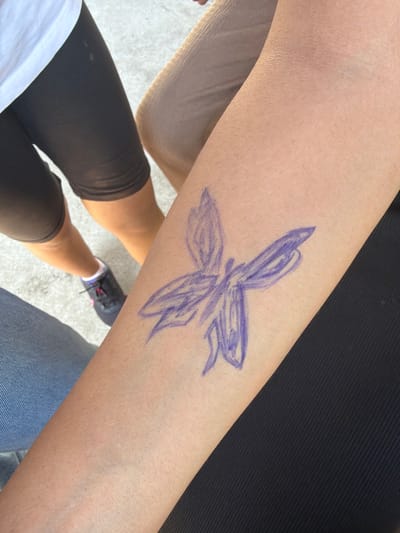 A close-up, eye-level shot shows a person's forearm with a purple, hand-drawn butterfly tattoo. The butterfly is centrally located on the forearm, with its wings spread. The forearm appears to be attached to a body wearing black clothing. In the background, out of focus, are legs and feet of other individuals, one wearing black athletic shorts and a white t-shirt, another wearing jeans, and a third person wearing beige. The setting appears to be outdoors, possibly on a paved surface like a sidewalk. The overall lighting suggests a sunny day. The image does not contain any information about the city of Dinkata, Bulgaria.