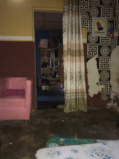 This image depicts an interior residential scene in Oba-Ile, Nigeria. The view is from one room looking into another through a doorway.
The foreground room features a two-tone wall on the left, with a yellow upper section separated by a white horizontal stripe from a dark red lower section. A pink, upholstered armchair with a pink cloth draped over it is positioned against this wall. The floor is a dark, unpolished surface, likely concrete, with some scattered debris and two patterned floor mats partially visible in the bottom foreground: one green with geometric shapes and another white with a floral design.
A wide doorway centrally frames the view into an adjacent room. A multi-patterned curtain, predominantly beige, brown, and white with decorative motifs, hangs vertically to the right of this doorway, partially obscuring the view. The room beyond the doorway has blue walls and contains an open shelving unit heavily loaded with miscellaneous items, including clothes, shoes, and containers. A small, multi-colored ball is visible on the floor of the second room.
The right wall of the foreground room is partially covered with black and white patterned wallpaper featuring a geometric Greek key design. A significant portion of this wallpaper or paint is peeling away, revealing the dark red substrate beneath. A framed picture depicting clasped praying hands and the word "ALHAMDOULLILLAH" hangs on this wall. Below the frame, two small, colorful balloons are partially visible near the floor. No individuals are present in the scene. The lighting is ambient and somewhat dim.