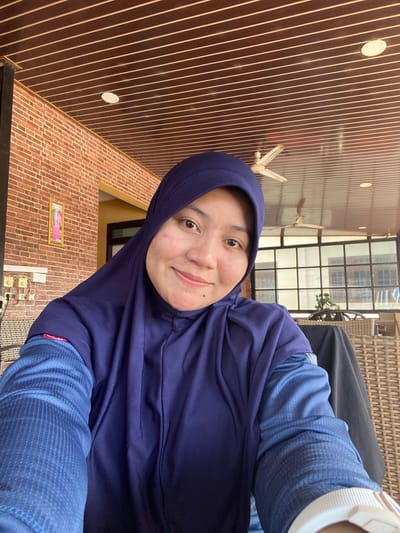 In an outdoor cafe setting, a woman wearing a deep purple hijab and a blue long-sleeved shirt smiles at the camera. She has light skin with some freckles and appears to be in her 20s or 30s. Behind her, a red brick wall and a slatted wooden ceiling are visible. A ceiling fan hangs from the ceiling, and large windows with dark frames look out onto another area with wicker furniture. The overall environment suggests a warm, casual atmosphere.