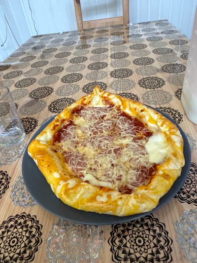 A pizza-like dish is centrally positioned on a dark grey plate, resting on a wooden table covered by a translucent plastic tablecloth patterned with repeating grey and silver mandala designs. The dish features a golden-brown, puffed crust, red sauce, and a topping of shredded white cheese and sliced pink meat, possibly ham or salami. To the left of the plate, an empty clear glass tumbler is present. A light-colored, opaque bottle or pitcher is partially visible on the right side of the table, behind the dish. In the background, a light-colored wooden chair back and a white vertically paneled wall are discernible. The scene is illuminated, located indoors in Temse, Belgium. No individuals are present.
