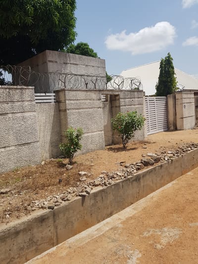 The image depicts a residential property in Palade, Nigeria. The foreground features a concrete drainage ditch running parallel to a sandy path. Beyond the ditch, a stone and concrete wall, topped with barbed wire, encloses the property. Several trees, including a large mango tree, are visible behind the wall, along with the corrugated metal roof of a building. A white metal gate is present in the wall, and a small bush with red flowers grows near the base of the wall. The sky is blue with scattered white clouds. There are no people visible in the image.