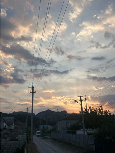 The image depicts a street scene in the city of Shchebetivka, Ukraine, under a dramatic, cloud-filled sky at dusk. The sky is illuminated with warm hues of orange, yellow, and pink from the setting sun, contrasting with darker, gray clouds. Several power lines stretch across the frame, converging towards utility poles that are visible throughout the scene.
In the foreground, a road leads into the distance, flanked by modest, low-rise buildings on either side. To the left, a house with a brick foundation and lighter-colored walls is partially visible. On the right, a fence separates properties from the street, and dense foliage from trees obscures some of the houses.
Further down the street, a white van is parked, and more houses with dark roofs are visible. In the background, a range of rugged, dark mountains rises against the twilight sky. The overall impression is a quiet, rural or semi-rural setting experiencing the transition from day to night.