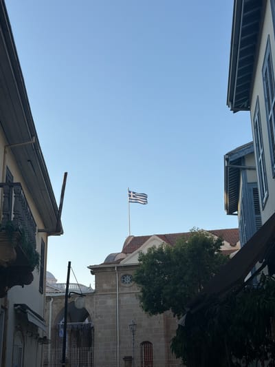 The image captures a low-angle view of buildings and the sky. A Greek flag is prominently displayed on a flagpole, fluttering in the breeze against a clear blue sky. The flag's design of white stripes on a blue background is clearly visible.

The surrounding architecture suggests an urban setting, possibly in Nicosia, Cyprus, as indicated by the architectural style. Buildings with tiled roofs and stone facades are visible, with elements like windows, shutters, and decorative architectural features. A lush green tree adds a touch of nature to the scene, with its branches reaching upwards. A lamppost is also present, an urban fixture.

The perspective is unusual, as if the viewer is looking up from a confined space between buildings, or perhaps the image is rotated. The time of day appears to be daytime, with the bright sky indicating good weather. No people are visible in the frame. The overall impression is one of a quiet, possibly historic, urban environment.