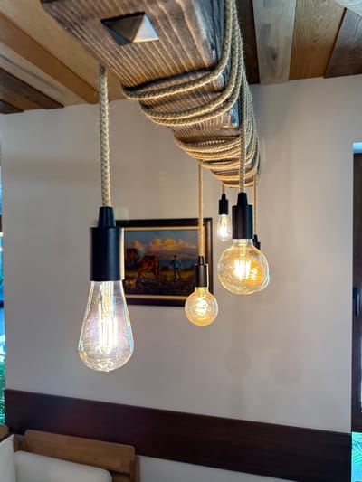This is a close-up, eye-level, medium shot of a rustic chandelier with exposed Edison bulbs hanging from a wooden beam in a room in Panichishte, Bulgaria. The chandelier is made of a rough-hewn wooden beam wrapped with thick rope. Five bulbs hang from black sockets by braided cords, each with a different shape: one pear-shaped, two round, and two smaller, elongated bulbs. The bulbs emit a warm, yellow glow. In the background, a framed painting depicts a horse and two figures in a landscape. The walls are painted a light, neutral color, and a dark wooden trim runs horizontally across the lower portion of the image. The ceiling is paneled with light-colored wood. The overall impression is one of a cozy, rustic ambiance.
