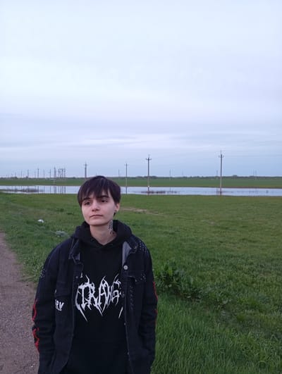 A young person with short dark hair, a tattoo on their neck, and a pierced lip stands on a dirt path. They are wearing a black hoodie with a white design and a black denim jacket with red details. The background is a flat, grassy landscape with utility poles stretching into the distance. There is a body of water reflecting the sky between the person and the utility poles. The scene is set in Azyakul', Russia.