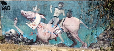The image is a wide-angle, eye-level, forensic photograph of a weathered mural on a turquoise, textured wall in Siero, Spain. The mural depicts a surreal, dark fantasy scene with several figures and elements.

In the center, a grotesque, pinkish-white, quadrupedal creature with exaggerated udders is ridden by a dismembered human figure. The rider, with a pale complexion and dark eyes, is headless, with the torso connecting to the lower body by a thin waist. The rider holds a dark stick across its shoulders, with a sack slung over one shoulder. A golden crown with white gems sits atop the creature's head. The creature itself appears to have internal organs exposed and is splattered with red and blue paint.

To the left of the creature, a pale, skeletal arm reaches upwards from a pile of skulls and bones. The skulls are stylized and appear to be canine or wolf-like.

To the right of the creature, the wall is partially obscured by dense, dry branches and leaves, casting shadows on the mural. The lower portion of the mural shows more skulls and skeletal remains, rendered in shades of black and gray.

The wall itself is heavily distressed, with peeling paint and visible textures, suggesting age and exposure to the elements. Some graffiti, including the letters "AHO" in black, is visible in the upper left corner. The ground in front of the mural is covered with dry grass and dirt. The lighting suggests it is daytime, with the sun casting strong shadows.