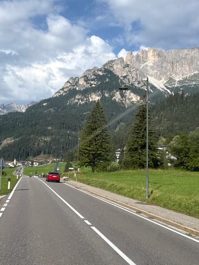The image shows a road leading towards the town of Mazzin in Italy, with a mountainous backdrop. A red car is visible on the road, approaching the viewer. The surrounding environment is characterized by lush green fields and dense pine forests on the slopes of the mountains. The sky is partly cloudy. In the distance, the town of Mazzin appears nestled among the hills, with buildings and a church steeple visible. The road itself is paved and marked with white lines. A lamppost stands on the right side of the road, and there are some trees along the roadside.