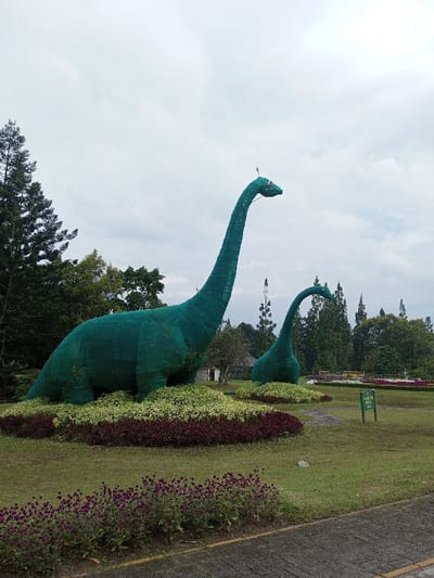Two green, life-size dinosaur topiaries stand in a park-like setting. The larger dinosaur towers over the smaller one, both with long necks and rounded bodies. They are surrounded by colorful flowerbeds with red, white, and yellow blooms. Beyond the dinosaurs, trees dot the landscape against an overcast sky. A paved path runs along the bottom of the frame, suggesting a walking area.