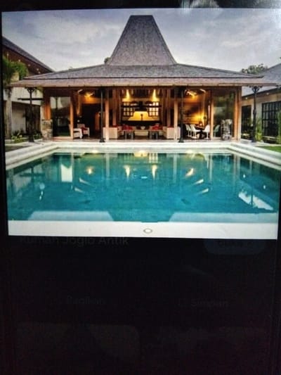 The image shows a luxurious outdoor living space with a swimming pool in the foreground and a traditional Indonesian-style building in the background. The building features a distinctive tiered roof, exposed wooden beams, and large glass windows that open to an indoor seating area. The swimming pool is a prominent feature, with clear blue water and a wide concrete deck surrounding it. The scene evokes a sense of tranquility and upscale resort living, likely in a tropical or subtropical climate. The text overlay at the bottom of the image, "Rumah Joglo Antik," translates to "Antique Joglo House," which is a traditional Javanese house. This suggests the location is likely in Java, Indonesia, or a place that features this architectural style.