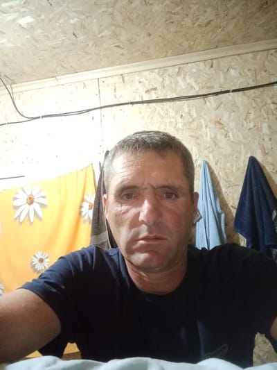 The image shows a man with short, graying hair and a fair complexion, looking directly at the camera. He is wearing a dark blue t-shirt. His facial features are prominent, with visible wrinkles on his forehead and around his eyes. The background consists of a wall made of plywood-like material, with electrical wires running across it. To the left of the man, a yellow curtain with white daisy patterns is visible. To the right, two towels are hanging on the wall; one is light blue, and the other is dark blue. The man is positioned in the foreground, occupying a significant portion of the frame. The overall scene suggests an indoor setting, possibly a room with a rustic or unfinished appearance. There is no information in the image to specifically pinpoint the location within Қарасу ауылдық округі, Kazakhstan.