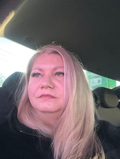 A female individual is depicted seated within a vehicle, observed in the city of Stary Chultem, Russia. She is positioned in what appears to be a passenger seat. Her long, light blonde hair exhibits a pinkish hue due to strong ambient light entering from the left. She is looking upwards and to her right. Her face shows defined eyelashes and lips with a reddish tint. She wears a dark, possibly black, zip-up garment with a visible metallic zipper pull. The vehicle's dark grey headliner is visible above, and a dark headrest is present behind and to her right. Through the vehicle's window, bright daylight is evident, along with faint horizontal lines resembling utility wires. In the background, a large green rectangular structure, possibly a building or sign, displays indistinct white graphical elements, including what may be a QR code.