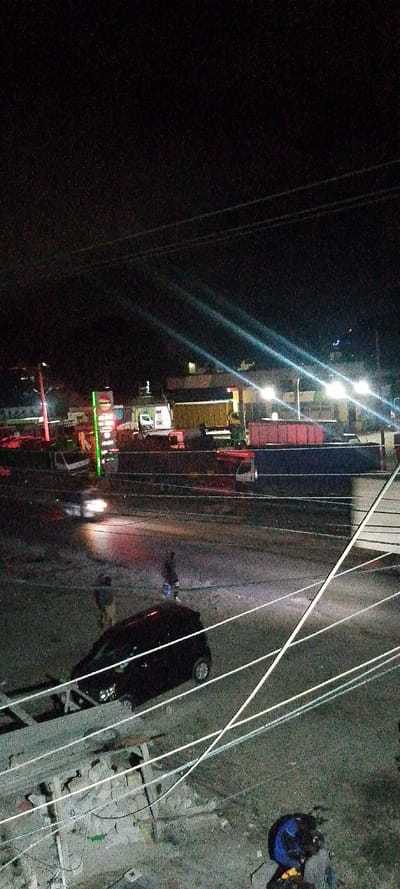 The image depicts an elevated night view of a street and commercial area in Frere Town ward, Kenya. The scene is illuminated by artificial light sources, creating prominent lens flares and visible digital noise.
In the immediate foreground, multiple white utility lines run diagonally across the frame, partially obscuring a paved or dirt surface. A dark-colored compact SUV or hatchback is parked or slowly moving on the right, with its rear section visible. Below and to the left of the vehicle, an unfinished concrete block structure is present. In the bottom right corner, a person is partially visible, wearing a dark top and a blue and black helmet or backpack.
The midground features a paved road where a vehicle with bright headlights is captured in motion, producing a light streak from left to right. Two male pedestrians are on the road surface: one is visible to the left of the moving vehicle's light, facing generally right; the other is to the right, holding a light-colored object (possibly a bucket) and facing generally left. Behind the road, a line of heavy vehicles is parked, including multiple semi-trailers loaded with red and dark blue shipping containers.
The background reveals a commercial complex featuring illuminated buildings and signs. A tall, vertical, green-lit sign, possibly for a petrol station, stands prominently on the left side of the background. Additional vans and trucks are visible around the buildings. The sky is dark and heavily affected by visual noise.