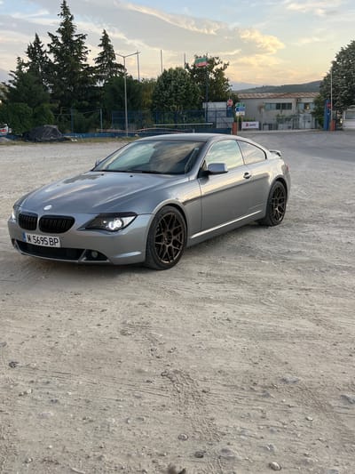 A silver BMW is parked on a dirt and gravel lot in Novo Delchevo, Bulgaria. The car is facing toward the right of the frame and angled slightly towards the viewer. The car's license plate reads "M 5695 BP". Behind the car are trees, a building, and a metal fence. The sky is visible in the background.