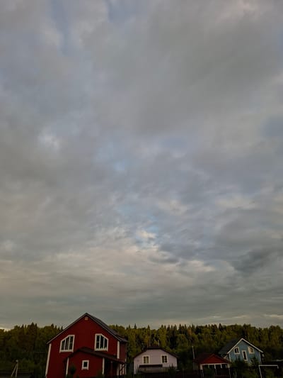 The image depicts a cluster of houses nestled amongst a dense forest. The houses are of varying colors and styles, including a red two-story house with white trim and distinctive arched windows, a light blue house with white trim, and a pale purple house. They appear to be residential properties. The scene is set against a dramatic sky filled with clouds, suggesting either dawn or dusk, or possibly overcast weather. The time of day is likely late afternoon or early evening, given the soft, diffused light. The location is identified as Ольшанка Кп, Russia, indicating a rural or semi-rural setting. The overall mood is peaceful and serene, with a sense of quiet habitation within nature. There are no people visible in the image, and no text is present. The focus is on the interplay between the man-made structures and the surrounding natural landscape.