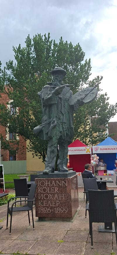 A bronze statue of the artist Johann Köler stands in the foreground, holding a palette and brushes. The statue is mounted on a granite pedestal inscribed with "JOHANN KÖLER MOXH KHLER 1826-1899". In the background, two tents are set up, one red and one blue, with vendors selling cotton candy and other goods. A man is seated at a table to the right, facing away from the camera. The scene is set in a paved outdoor area with trees and buildings visible behind the tents. The sky is overcast. The statue is located in the city of Viljandi, Estonia.