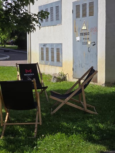 This is a medium shot, eye-level, outdoor photograph taken on a sunny day. In the foreground, on a patch of green grass with some small white flowers, are three wooden and black fabric deck chairs. One chair in the center has a red and white logo with the word "МОЗАЙКА" (MOSAIKA) and "espresso bar" printed on it. The chair in the foreground on the left is solid black. A third chair is partially visible on the right. Behind the chairs is a weathered, light gray building with several metal grates and a metal door. The door has a yellow triangle with a lightning bolt symbol and Ukrainian text indicating it's an electrical hazard, along with other markings. The building is stained with yellow and has some graffiti. To the left of the building, part of a leafy green tree branches into the frame, casting shadows. In the background, a paved area and some trees are visible, suggesting an urban or suburban setting. This scene is located in Petropavlivska Borshchahivka, Ukraine.