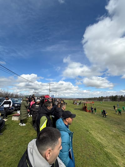 The image depicts an outdoor scene in Petarnitsa, Bulgaria, featuring a football match observed by a crowd of spectators under a bright, partly cloudy sky. In the foreground, the backs and heads of several male spectators are visible, positioned on a grassy embankment overlooking the field. A man in a light blue puffer jacket and a black baseball cap is prominent on the right, his gaze directed towards the field.

The midground shows a large gathering of individuals, predominantly adults, standing on a grassy area adjacent to a parking lot filled with dark-colored vehicles. Overhead, multiple black power lines traverse horizontally. Further into the midground, on a descending grassy slope, two open-sided shelters with red-brown roofs are situated near the football pitch.

On the green grass of the football field in the background, multiple individuals are present, appearing to be players and officials. Several players are distinctly visible in green jerseys, while others wear yellow/black or blue. A white goal frame stands at the far end of the field. A line of deciduous trees forms the distant boundary of the playing area, beyond which a hazy horizon is discernible. The overall lighting suggests a sunny day.