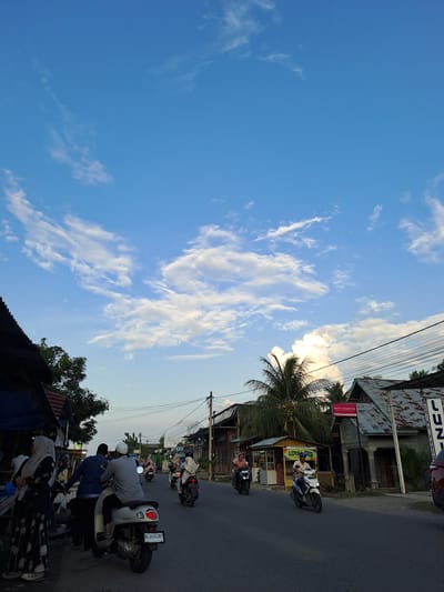 The image depicts a street scene in Cottarum Tunong, Indonesia, under a bright blue sky with scattered white clouds. The road is paved and lined with buildings, some of which appear to be shops or residences with corrugated metal roofs. A few palm trees are visible, suggesting a tropical or subtropical climate. Several people are on the street, mostly riding motorcycles.
On the left side of the road, a person wearing a hijab and a patterned dress is standing near a stall or shop. Two men are on a scooter in the foreground, with one driving and the other seated behind. Further down the road, other people on motorcycles are visible. One motorcycle has the license plate "BL 3404 ZBT". A red car is partially visible on the far right of the frame. Signs for businesses such as "LOYA" and "KITTY COSMETIK" are present on the buildings. Power lines stretch across the scene, supported by utility poles. The overall impression is a bustling street during daylight hours.