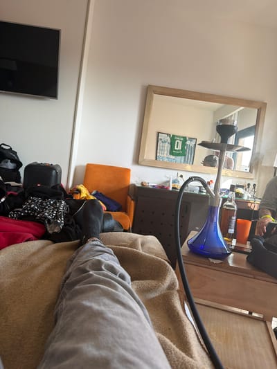 The image shows an indoor scene, likely within a residence, containing various objects and potential activities. A person is visible, sitting with their leg extended, dressed in grey jeans and black socks. Near them is a tan-colored blanket or throw draped over the furniture. A hookah pipe, with a blue glass base and black hose, is prominent. It sits on a wooden table cluttered with items including liquor bottles, snacks, and an orange cup. A reflection in a mirror on the wall displays the hookah, surrounding objects, and possibly a person's face in the background. Below the mirror, a framed jersey with the number 10 is visible. A flat-screen television is mounted on the wall further down. Other items include an orange chair, a black suitcase, a speaker, and assorted clothes scattered on the floor. The overall impression is one of casual disarray. Location cannot be verified as Punta Umbría, Spain, based solely on image analysis.