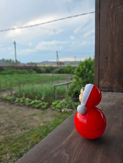 A close-up shot shows a red plastic toy, likely a Santa Claus figurine, resting on a weathered wooden surface. The toy is positioned to the right of the frame, with its rounded red body and a white trim suggesting a hat. A glimpse of a small, light-skinned face is visible beneath the white trim. The background is blurred, revealing a green garden area with rows of plants under a bright, hazy sky. The sky appears to be a light blue with streaks of white clouds, and the sun is visible, indicating daylight. The overall scene suggests an outdoor setting, possibly a backyard or a rural area. The wooden surface has a rough texture and appears to be part of a railing or a deck. The focus is primarily on the toy, while the background provides a sense of place. The image is oriented sideways.