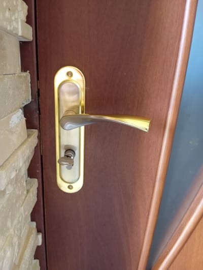 A brass door handle with a silver lever and a key inserted into the lock is visible on a dark brown wooden door. The door is positioned next to a wall constructed of light-colored stacked stone. The scene is likely an exterior shot of a residential or commercial building in Hlynytsi, Ukraine.