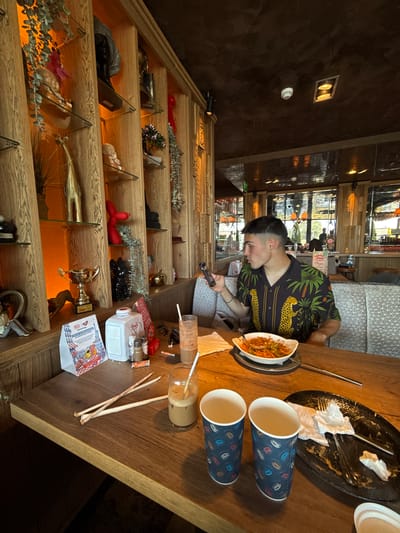 The image captures an indoor scene, likely a restaurant or culinary center in Radinovo, Bulgaria, oriented vertically (rotated 90 degrees counter-clockwise). On the right, a young man with a short, faded haircut is seated at a wooden table, engrossed in his smartphone. He wears a dark short-sleeved shirt patterned with green palm trees and yellow leopards.
On the table in front of him are the remnants of a meal in an oblong white dish, a dirty black plate with used napkins, and two blue paper cups with a colorful leaf pattern. An empty glass jar with a straw suggests a finished beverage, and various small items like sachets and a white bottle (possibly labeled "Happy Original") are also present. The background behind the man shows large windows with reflections of an outdoor scene, suggesting it is daytime.
To the left, a multi-tiered wooden shelf unit extends vertically, filled with a diverse collection of decorative objects. These include a golden trophy, small statues (such as a baby doll figure), artificial plants, bottles, a red balloon animal, and a black panther figurine. The shelves are warmly lit from below. A prominent white sign with a red border on one shelf clearly reads "РАДИНОВО" (Radinovo), "КУЛИНАРЕН ЦЕНТЪР" (Culinary Center), and "BULGARIA", confirming the location as a culinary center in Radinovo. Another small card visible on a shelf has "HAPPY CLUB" written on it. The walls are a dark, textured material, and ceiling lights are visible. The overall ambiance appears casual and well-decorated.