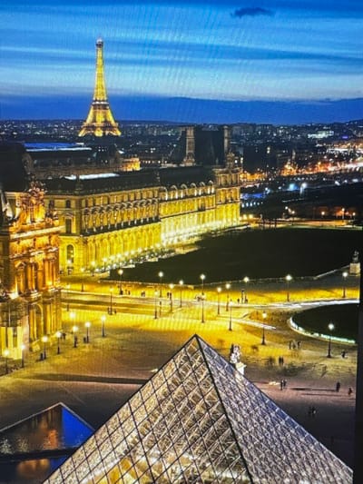 This is a rotated, nighttime aerial view of Paris, France. The primary subjects are iconic landmarks illuminated by artificial lights. The Eiffel Tower is visible on the left, standing tall against a bright blue sky with some clouds. To its right, a long, grand building, likely the Louvre Museum, stretches across the frame, its facade lined with numerous lights. In the foreground, a large glass pyramid structure, also part of the Louvre, is prominently featured, reflecting the surrounding lights. The scene depicts a city at night, with the lights of buildings and streets creating a warm, golden glow. There are no people or specific activities clearly discernible due to the distance and lighting. The location context provided (Kuvshinovo, Russia) is inconsistent with the visual content, which clearly shows Parisian landmarks.