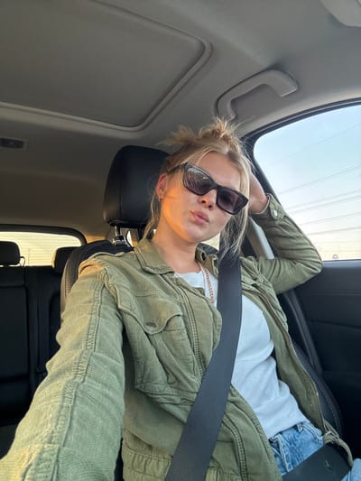 A woman is depicted seated inside a vehicle, identified as being in Kafr Kasim, Israel. She is positioned in the front passenger seat, secured by a black seatbelt across her chest and shoulder. Her blonde hair is styled in a messy bun. She wears dark, cat-eye style sunglasses, a white t-shirt, a light green utility-style jacket with multiple pockets and buttoned flaps, and blue acid-wash jeans. Her right arm is extended, holding a device to capture the image, while her left hand rests behind her head. Her lips are slightly puckered. Warm, golden light illuminates the left side of her face and clothing, indicating a low sun angle. The car's interior features include dark upholstery, a sunroof, and an overhead grab handle. Through the windows, a clear sky is visible, transitioning from blue to a lighter hue, with faint outlines of utility lines or poles in the distance.