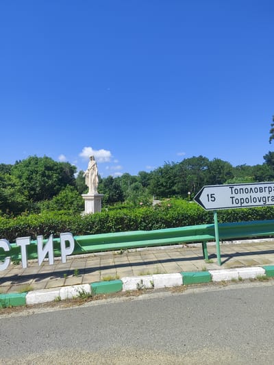 The image depicts a bright, clear day with a vibrant blue sky and a crescent moon visible. In the foreground, a green metal guardrail runs horizontally across the frame. To the left, large white three-dimensional letters spell out "СТИР," partially obscured by the guardrail. The ground beneath the letters is paved with square concrete tiles, bordered by a curb painted with alternating green and white sections.
Behind the guardrail and letters, a manicured hedge forms a dense green backdrop. To the left, a white statue of a robed figure stands on a pedestal amidst the foliage. The statue appears to be a historical or religious monument. A directional signpost is visible to the right, indicating "15" and "Тополовгра Тороlovгра" with an arrow pointing to the right, suggesting a distance and a destination likely within or near Golyam Manastir, Bulgaria. The background is filled with lush green trees, indicative of a park or natural setting. The overall scene suggests a roadside view in a scenic location.