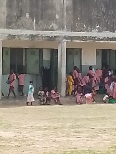 The image depicts a crowded outdoor scene in Igbo Ijaiye, Nigeria, showing numerous children gathered in front of a two-story concrete building. The building's upper facade is weathered and discolored, while the lower level features multiple rectangular doorways or windows with dark, possibly green, panels. A raised concrete platform or step runs along the base of the building, serving as a seating area for some individuals. The ground in the foreground is an unpaved, light-colored, dry dirt or dusty area, with minimal visible vegetation.

Approximately twenty-five to thirty individuals, predominantly children aged roughly 5-12, are visible. Most children are dressed in similar light-red, pink, or maroon-colored short-sleeved shirts and shorts or skirts, consistent with school uniforms. Notable exceptions include one child on the far right wearing a yellow long-sleeved outfit and another child in the mid-left foreground wearing a light blue/green top and a cream-colored skirt. One boy seated on the left wears a camouflage-patterned top and shorts. An adult-like figure or older child in a dark blue/black shirt is seen from the back among a cluster of children on the right.

Actions include children standing, walking, and sitting. On the far left, two boys in pink/maroon uniforms are standing, with one appearing to walk towards the left. In the mid-left, a small group of children, including the one in the light blue/green and cream outfit, are seated on the concrete platform. On the right side, a larger group of children is densely clustered around an entrance, some appearing to be in motion or interacting, suggesting a period of recess or transition between classes. No specific objects beyond clothing are clearly identifiable due to the low resolution of the image. The scene is illuminated by natural daylight, casting no distinct shadows.