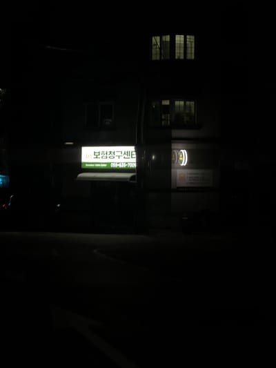 The image depicts a nighttime scene in a dark urban environment, likely in Geoje-si, South Korea, as indicated by the location context. The primary subject is a brightly lit green and white sign on the side of a building. The sign displays Korean text and what appears to be a phone number.
The sign's text is in Korean, with the larger characters reading "보험청구센터" which translates to "Insurance Claim Center". Below this, smaller text reads "Insurance Claims Center" in English, and then a phone number "055-635-7009" is clearly visible. There is also a small circular sign with a green ring and white center, possibly a logo.
The building itself is dark, with several windows visible. Some windows have internal lights on, showing a grid-like pattern within. The overall atmosphere is dim and quiet, typical of a late hour in a commercial or residential area. The perspective suggests the photo was taken from the street level, looking up at the building. There are no people clearly visible, and the focus is on the illuminated sign which stands out against the darkness. The weather is not discernible from the image. The scene evokes a sense of a business operating or being represented in the night.