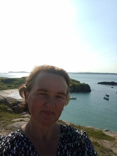 A woman takes a selfie from a rocky, grassy incline overlooking a bay in Carnboy, Ireland. The woman, facing forward, has light brown hair pulled back and is wearing a black and white patterned dress. The bay, which appears calm, contains several small boats anchored in the turquoise water. In the distance, a sandy beach can be seen with a few figures walking. Islands and hills form the horizon under a bright, clear sky.