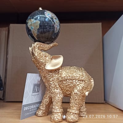 A golden, ornate elephant figurine is displayed on a wooden surface, holding a small globe on its raised trunk. The elephant is intricately detailed with floral patterns. A white tag with the text "AD TREND", "DECORAZIO", "IN RESINA", and "CON MAPPAMO" hangs from its trunk. In the background, cardboard boxes and a shelf are visible. The image also includes a timestamp "24.07.2026 10:57" in the lower right corner. The scene appears to be an indoor setting, likely a shop or a home, possibly in Modugno, Italy, though this cannot be definitively confirmed from the image itself.