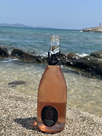 A bottle of rosé wine sits on a pebbled beach with the turquoise water of the sea and a rocky shoreline in the background. The bottle is condensation-covered, indicating it is chilled. The label on the bottle reads "TERRE DI TALOS PUGLIA VINO SPUMANTE EXTRA DRY ROSE." The scene appears to be located in Castiadas, Italy, given the context of the Puglia wine and the coastal scenery often found in that region. The sky is clear and blue, and the sun is shining.