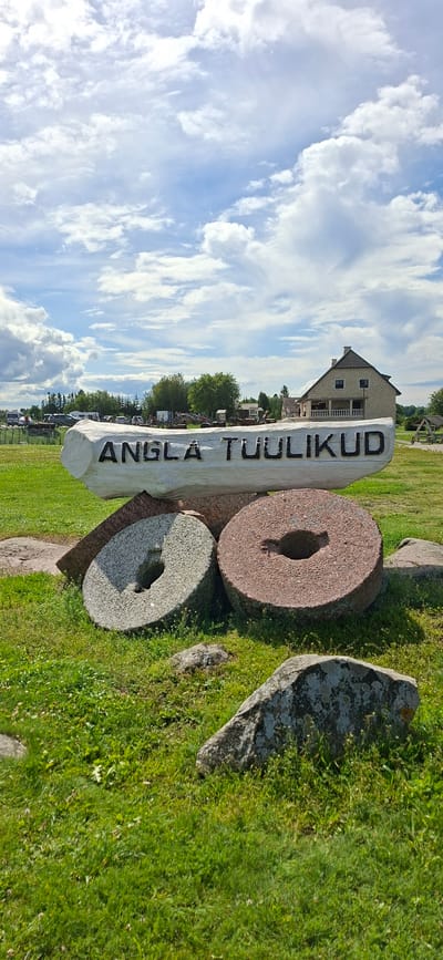 In Angla, Estonia, a white wooden sign carved with the words "ANGLA TUULIKUD" stands horizontally, supported by large, round millstones. One millstone, gray and speckled, rests on its side in front of the sign, while another, reddish-brown, is propped upright to its right. The scene is set outdoors on a grassy area, with a few scattered rocks and patches of wildflowers. In the background, a stone building with a dark roof and multiple windows is visible, along with trees and a cloudy sky. A fence and several vehicles, including what appear to be RVs, are also present in the distance.